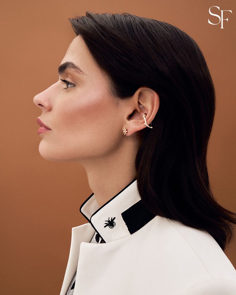 Profile portrait of a woman wearing gold earrings and a white jacket against a brown backdrop in a studio-lit environment.