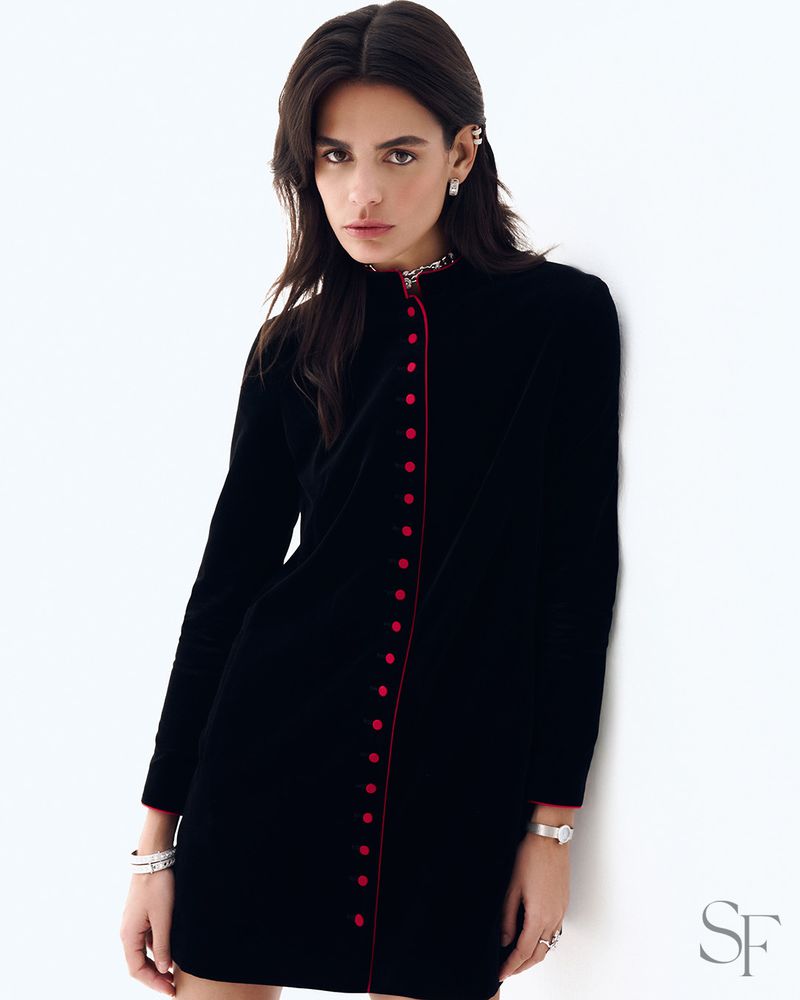 A woman with dark hair wearing a black long-sleeve buttoned mini dress with red trim against a white background.