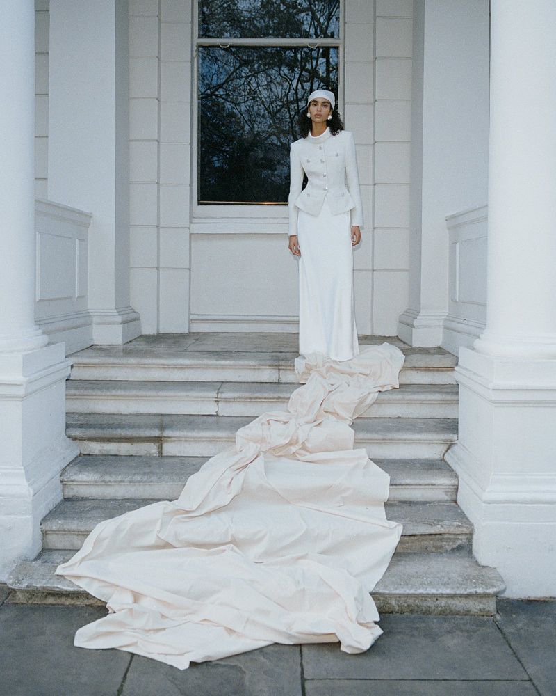 Elegant woman in white suit stands on stairs with flowing fabric. Outdoor fashion portrait with natural light and architectural backdrop.