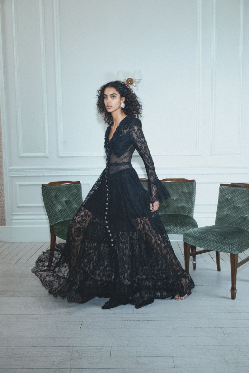 Model Noemi Kassa poses in a full-length black sheer lace tiered gown standing in front of green tufted chairs.