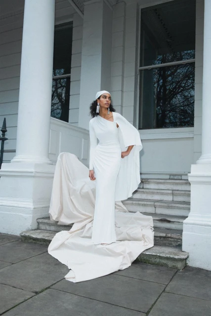 An adult female in a white gown poses on stone steps outside a building with white columns and windowed openings on a cloudy day.