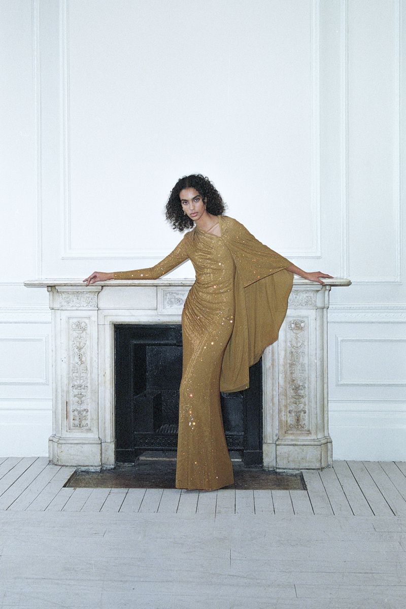 Fashionable woman in a gold sequin gown poses elegantly by a fireplace in a photo studio, exuding sophistication and style in a minimalist setting.