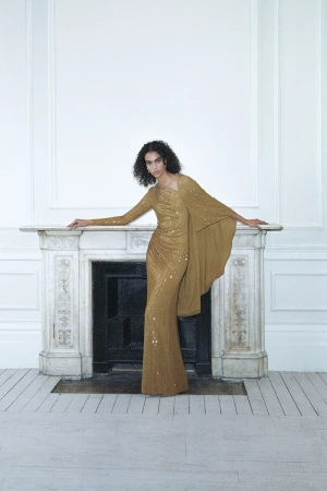 Fashionable woman in a gold sequin gown poses elegantly by a fireplace in a photo studio, exuding sophistication and style in a minimalist setting.