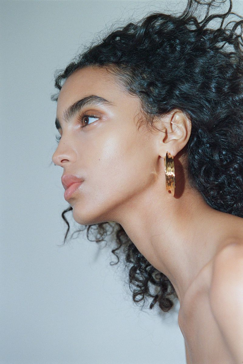 Side profile portrait of model Noemi Kassa with dark curly hair wearing a textured gold hoop earring against a pale studio backdrop.