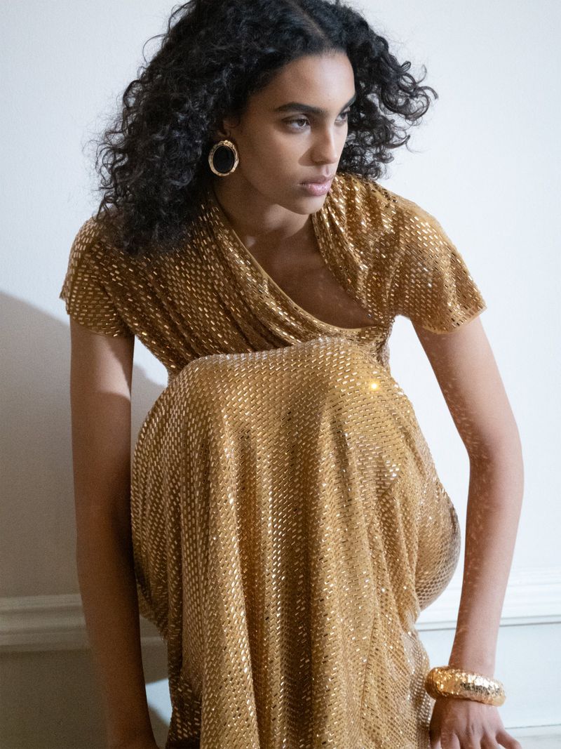 A woman poses in a glittering gold, draped gown. She wears gold hoop earrings and a bangle bracelet, styled for fashion or editorial.