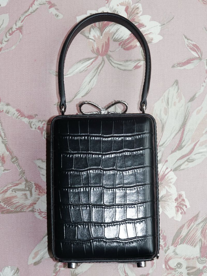 Black clutch handbag with a rectangular body and a curved handle, adorned with a sparkly bow detail against a floral wallpaper background.