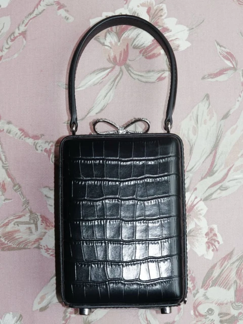 Black clutch handbag with a rectangular body and a curved handle, adorned with a sparkly bow detail against a floral wallpaper background.