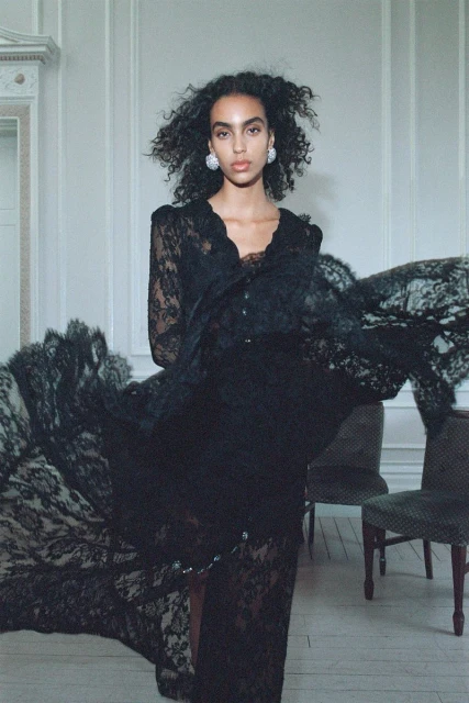 A woman poses indoors wearing a long-sleeved black lace dress, statement earrings, and neutral makeup, with white walls in the background.