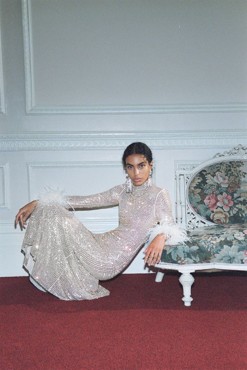 Elegant woman in a sparkling gown with feather accents sits on a floral sofa, posing indoors against a pale blue paneled wall.