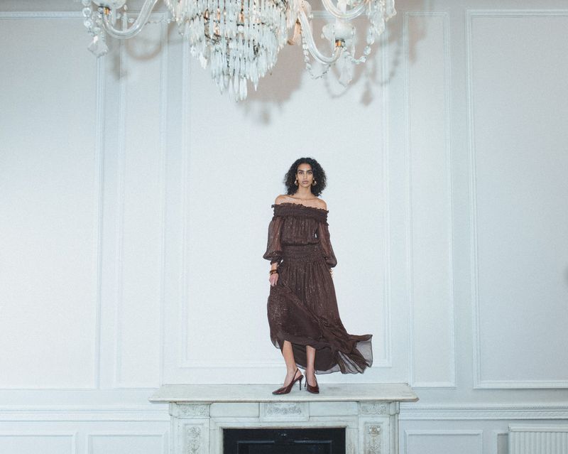 Woman in a brown off-the-shoulder dress stands on a white mantelpiece in an ornate room with a crystal chandelier overhead.