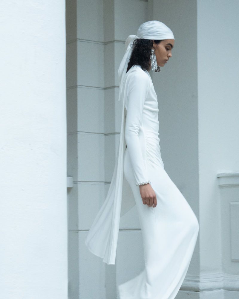 Woman in flowing white dress and headscarf poses against a white pillar, showcasing fashion with draped fabric and elegant design.