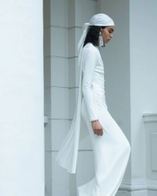 Woman in flowing white dress and headscarf poses against a white pillar, showcasing fashion with draped fabric and elegant design.