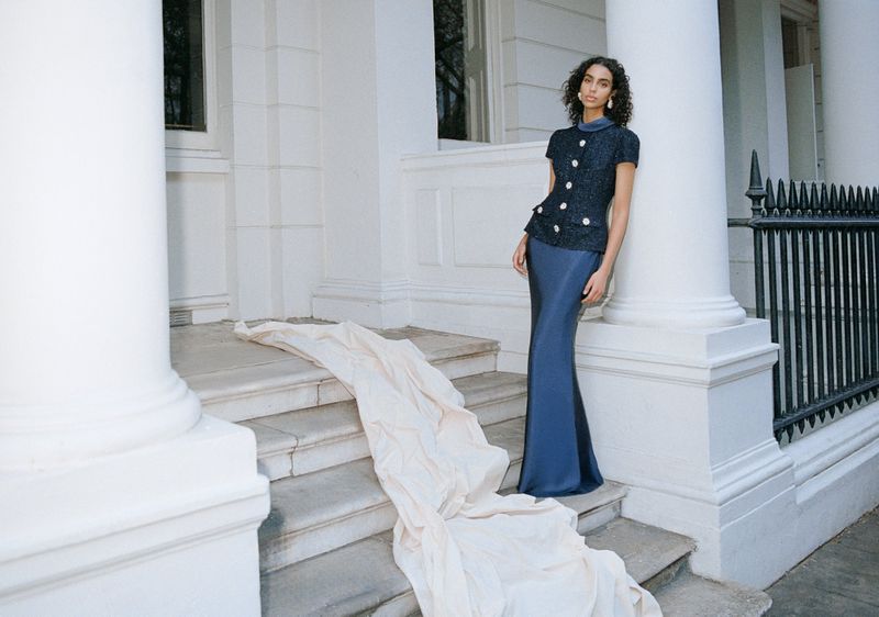 Woman in a navy top and long skirt poses on marble steps near a white stone pillar, next to a cream-colored cloth draped on stairs.