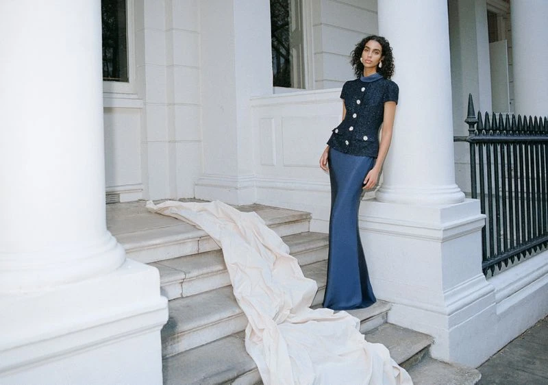Woman in a navy top and long skirt poses on marble steps near a white stone pillar, next to a cream-colored cloth draped on stairs.