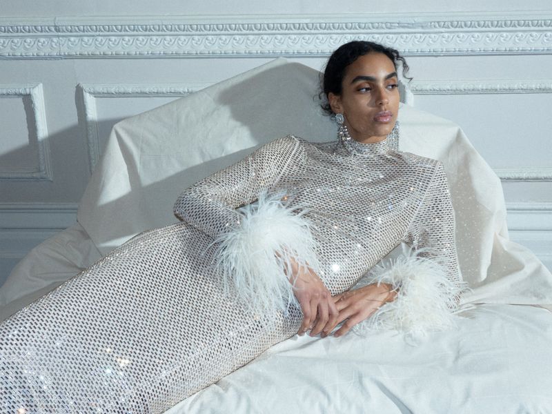Elegant woman in a shimmering dress with feather trim reclining on a bed against a white wall for a sophisticated fashion advertisement.