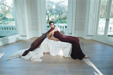Model Noemi Kassa reclines on a fabric-covered plinth in a strapless burgundy gown with a long flowing scarf beside large sunlit windows.
