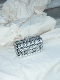 A rectangular black evening clutch encrusted with teardrop crystals and a bow clasp resting on draped cream fabric.