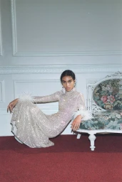 Model in a crystal-embellished gown with feather cuffs posing next to an antique floral settee