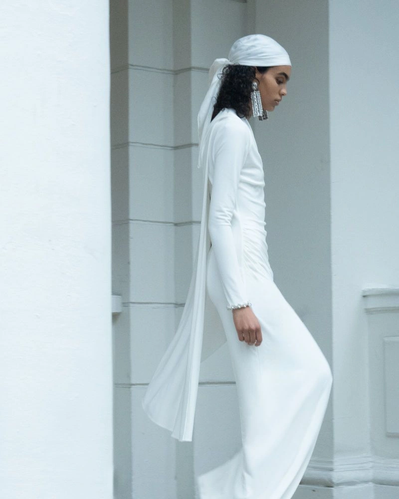 Model in a long white dress and matching headscarf standing against a light architectural background.