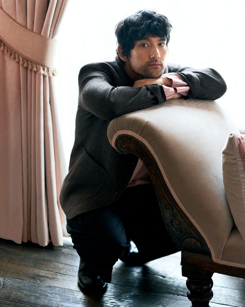 A man in a blazer poses indoors with a contemplative expression, leaning against an ornate lounge chair next to a curtained window.