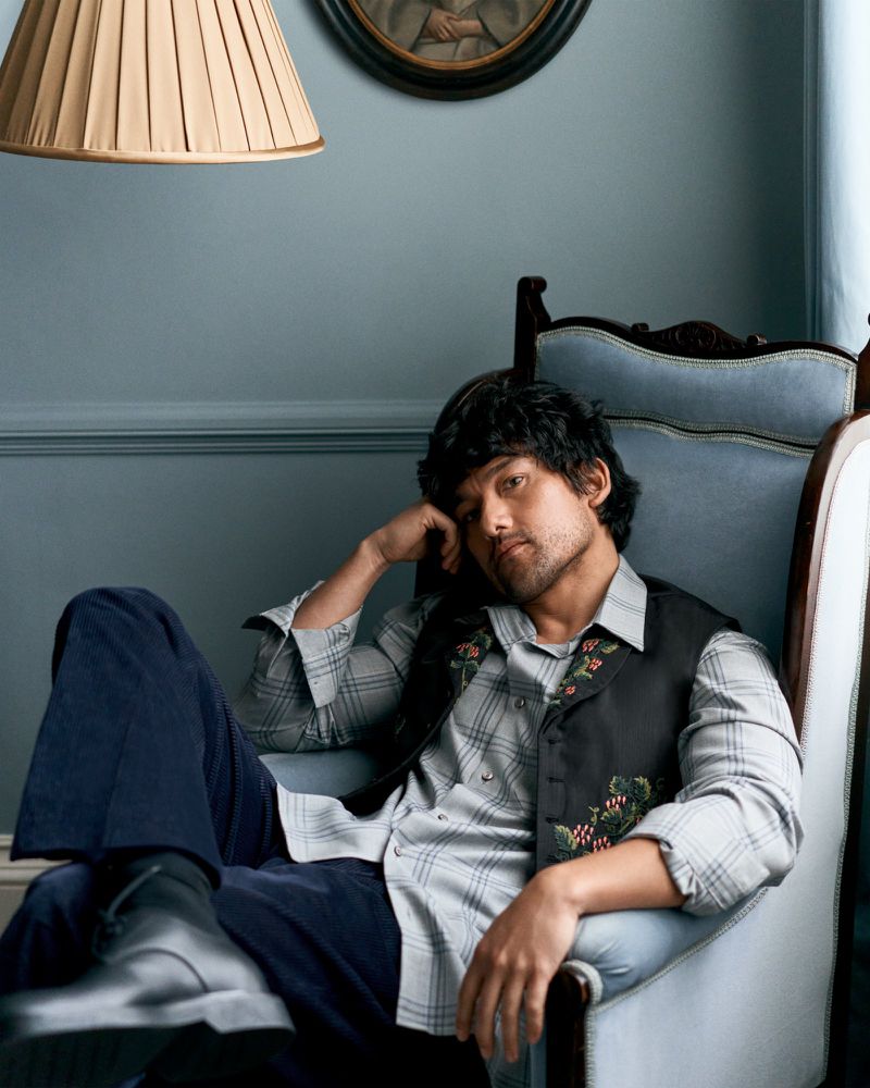 Actor Will Sharpe seated in a vintage blue armchair wearing an embroidered black vest and plaid shirt.