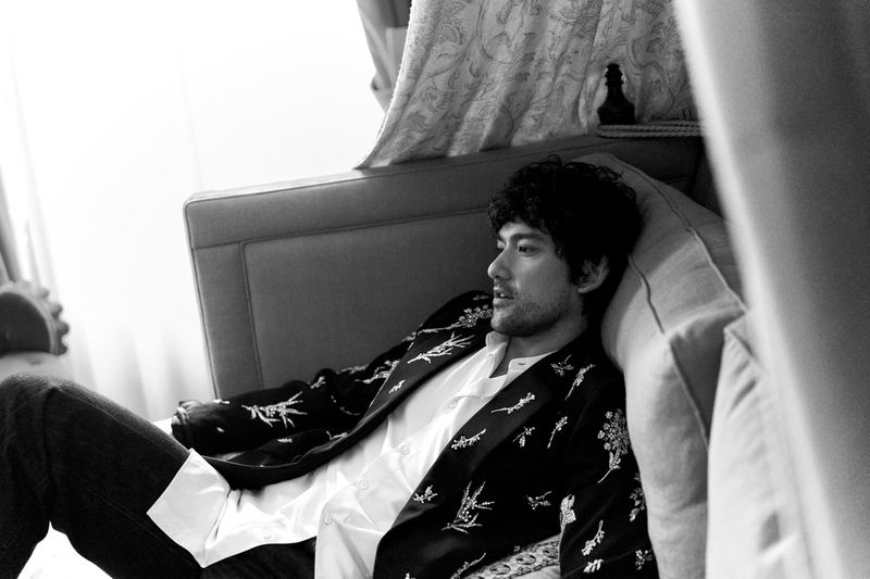 Monochrome portrait of a reclining adult male with dark hair, wearing a patterned blazer and white shirt, in a naturally lit interior.