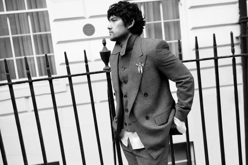 Monochrome portrait of an adult male in a grey suit standing outdoors next to an iron fence and a light-colored building with windows.