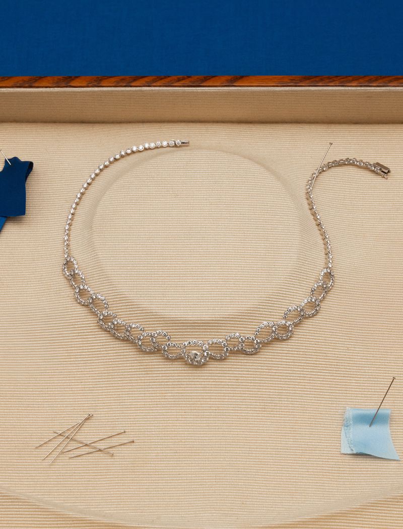 Elegant diamond tennis necklace on a light beige surface with fabric scraps and sewing pins against a blue background.