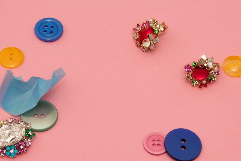 Flat lay of colorful buttons and gemstone brooches scattered on a pastel pink backdrop, creating a playful and decorative arrangement.