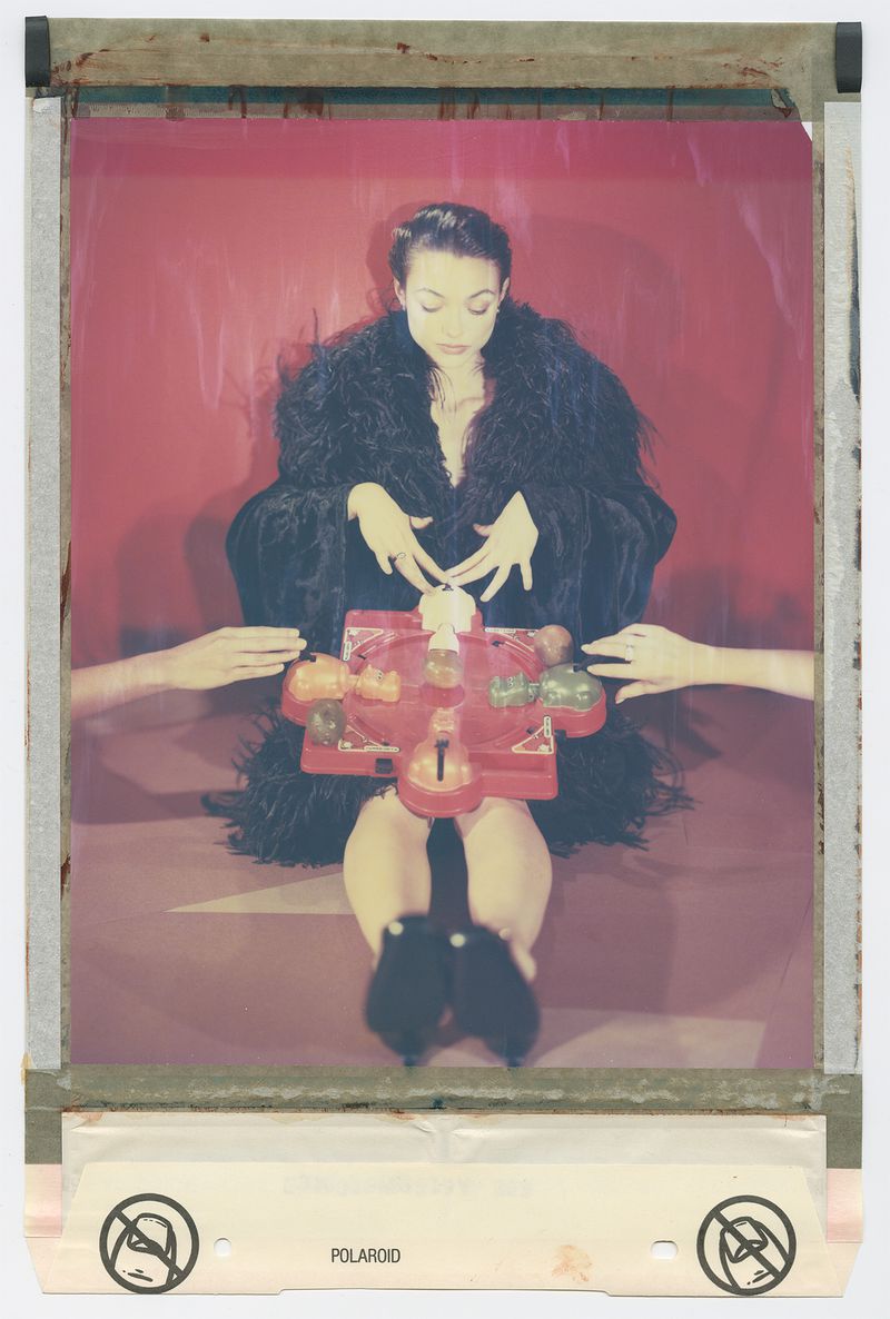 Woman in a feathered robe sits behind a red game with hands around it, captured in a Polaroid-style image against a red backdrop.