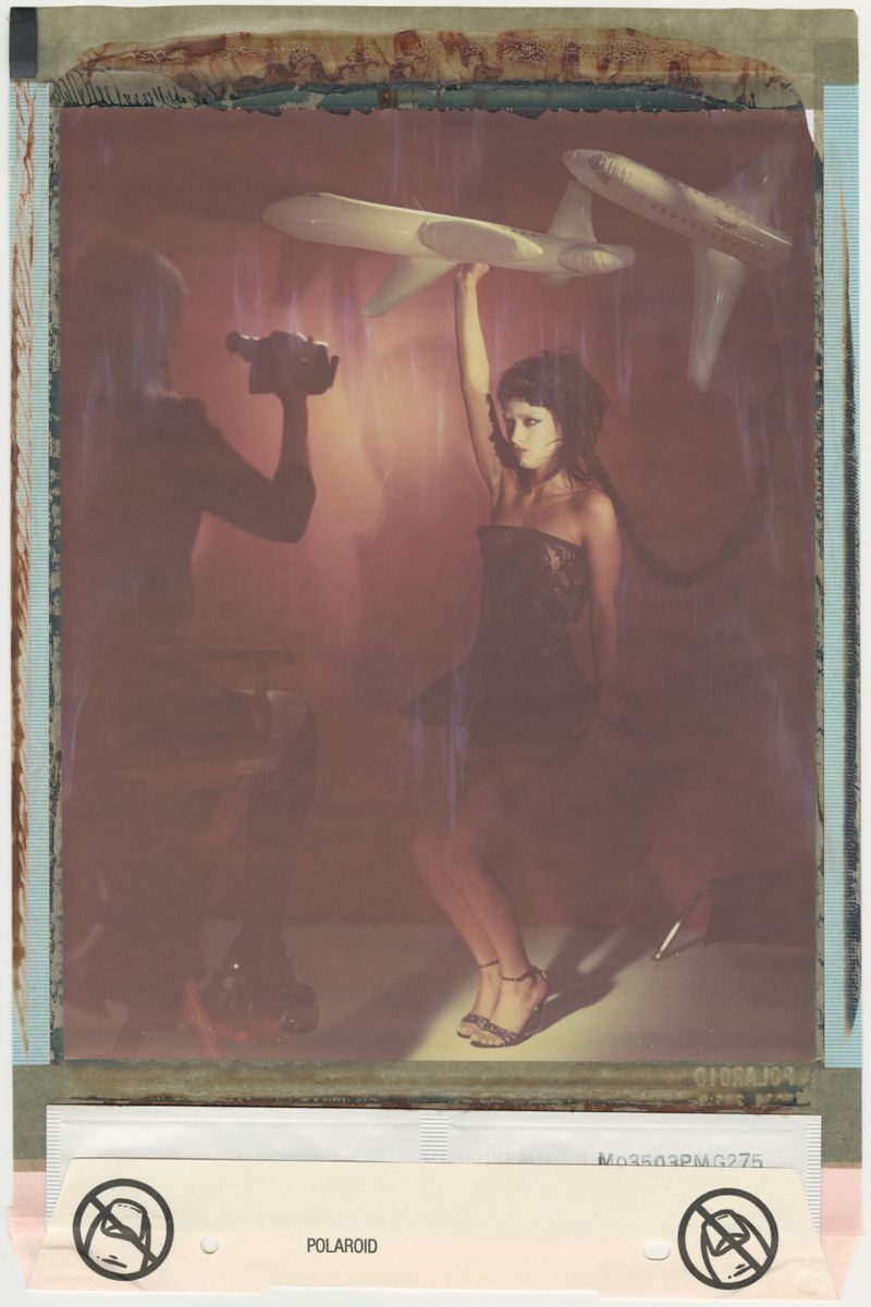 Woman in dress holds model airplane, while person holding a camera stands in the background in a vintage Polaroid photograph.
