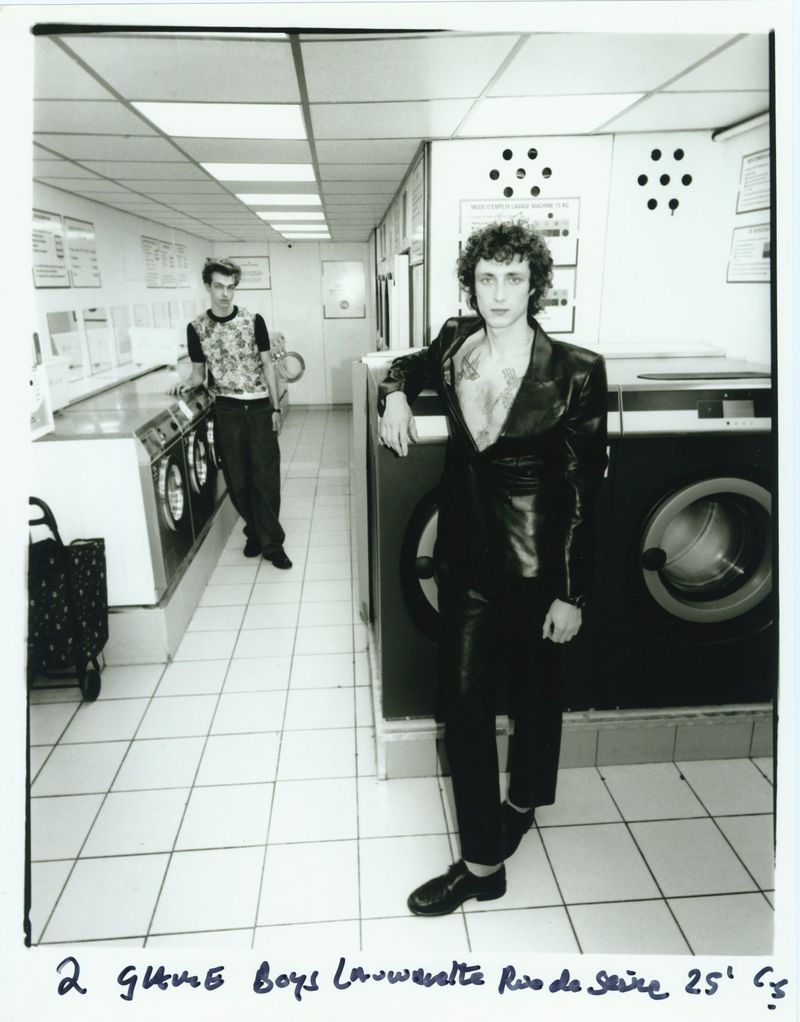 Black and white medium shot of two men in a laundromat, one in a shiny black jacket leaning against a washing machine.