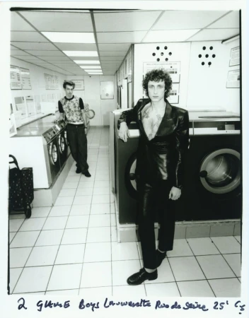 Black and white medium shot of two men in a laundromat, one in a shiny black jacket leaning against a washing machine.
