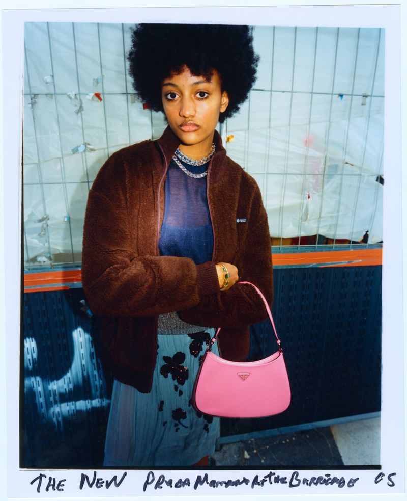 A young woman with an afro poses with a pink Prada handbag in front of an urban background with graffiti and fencing. Fashion shoot.