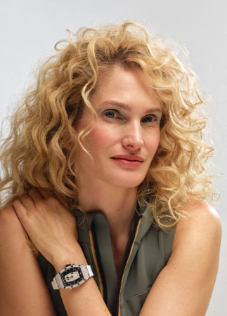 Close-up studio shot of a blonde adult woman wearing a sleeveless top and a white watch, with her arms crossed and a neutral expression.