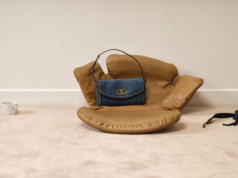Denim Valentino handbag on a tan leather chair with off-white carpet and walls in a studio setting, showcasing fashion accessory.