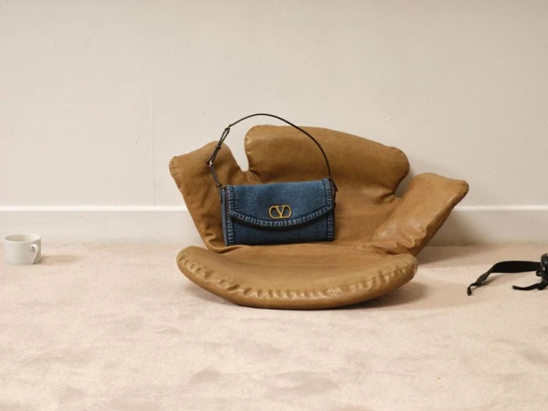 Denim Valentino handbag on a tan leather chair with off-white carpet and walls in a studio setting, showcasing fashion accessory.