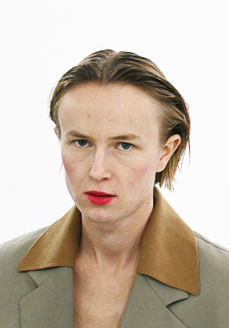 Close-up portrait of a person with slicked-back hair, wearing a beige blazer with a tan collar, set against a clean, bright white background.