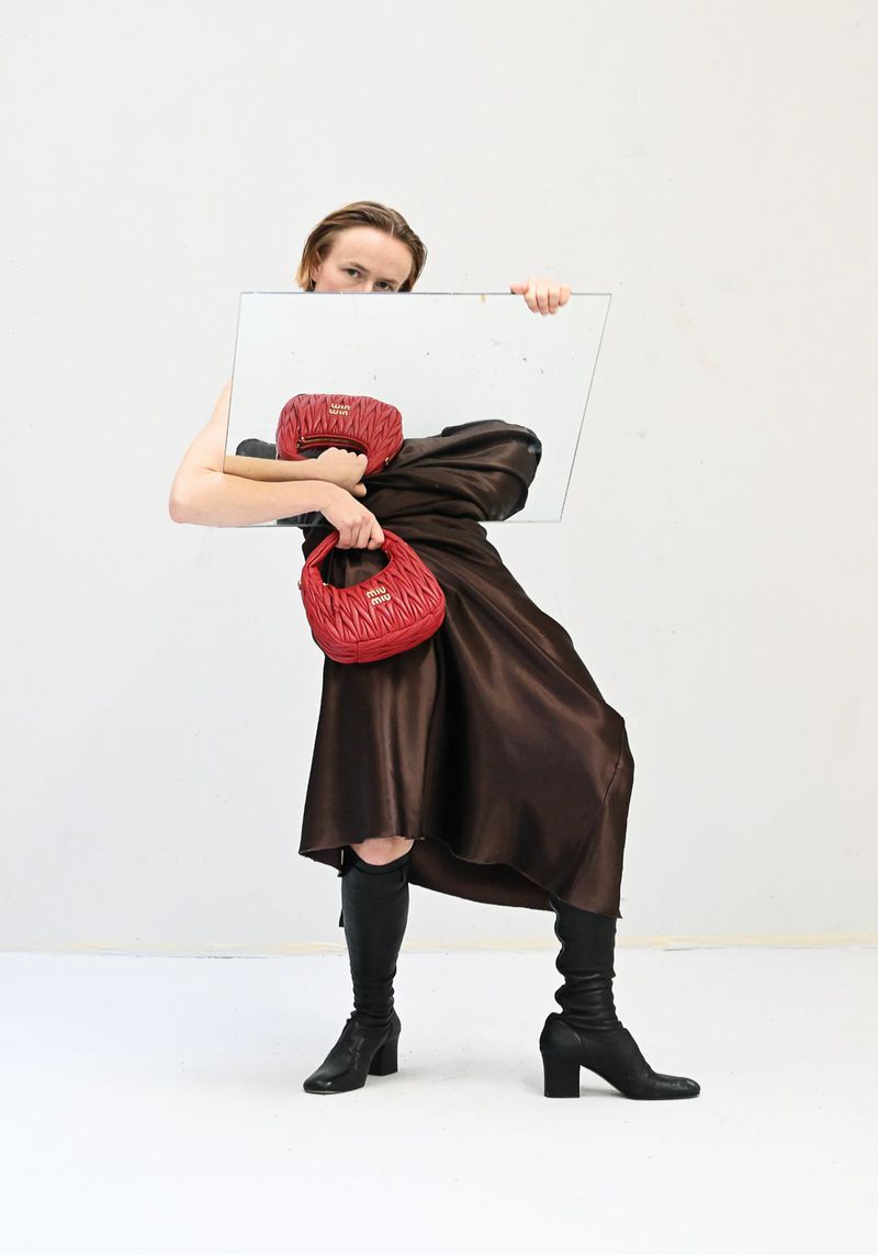 Woman strikes a pose with a red purse and mirror in a studio. She wears black boots and a brown dress against a white backdrop.