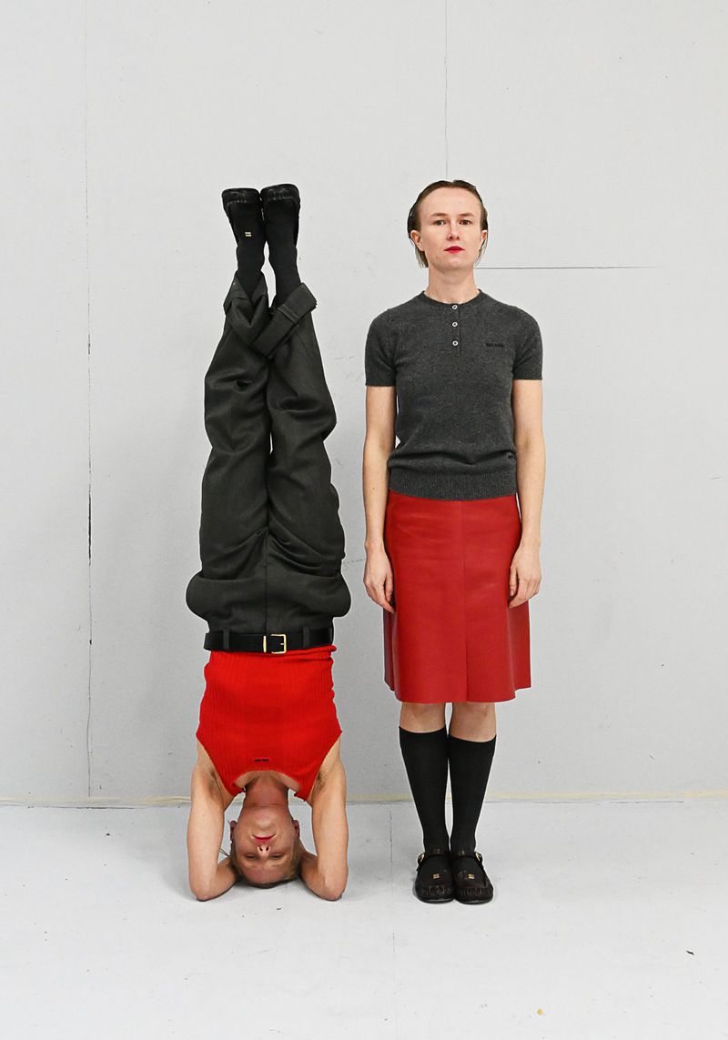 Two women in a studio setting, one in a headstand wearing a red tank top and dark pants, another standing in a gray top and red skirt.