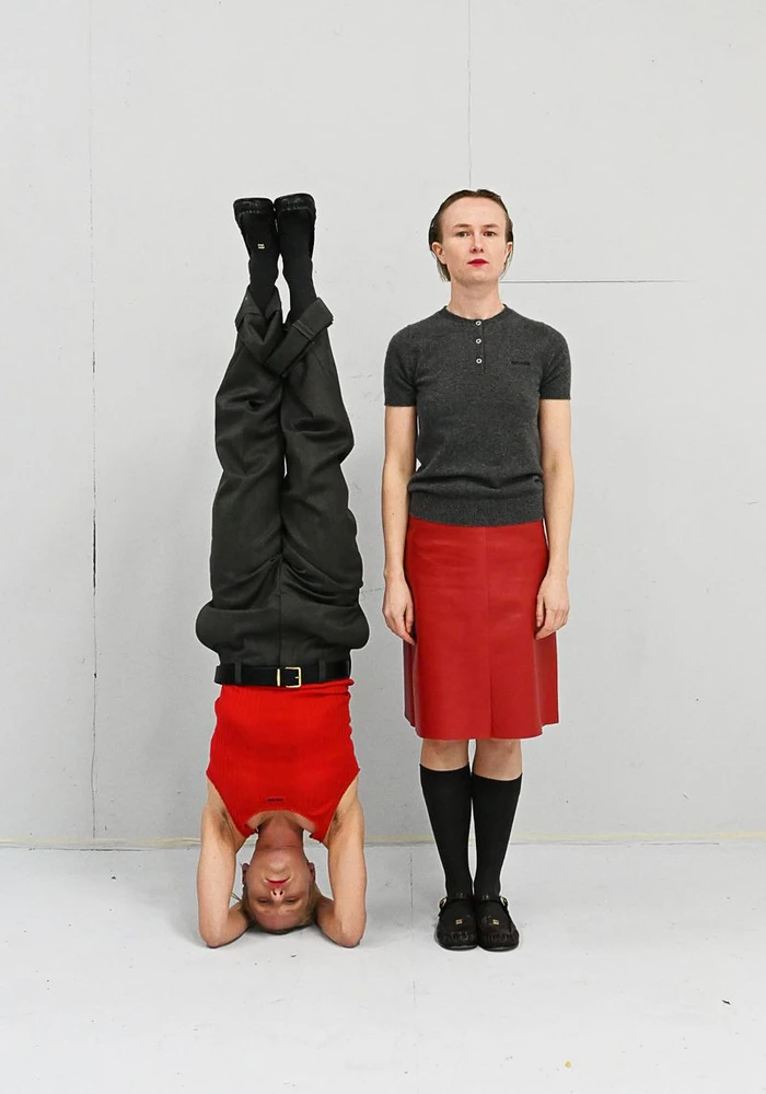 Two models posed against a grey wall, one standing upright in a red skirt and grey top, the other upside down in a headstand.