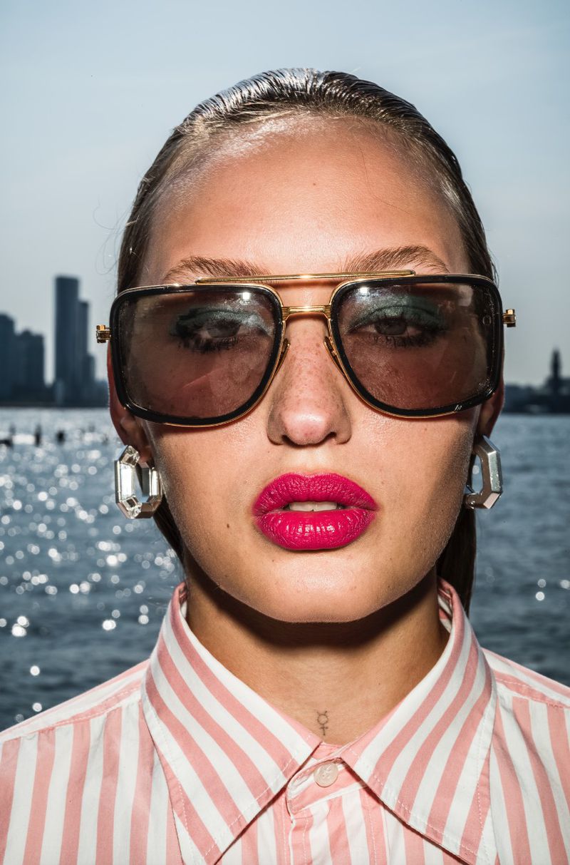 Close-up portrait of a woman wearing oversized aviator sunglasses, pink lipstick, and a pink striped collared shirt by water.