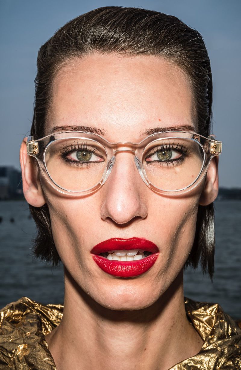 Close-up portrait of a woman with red lipstick and clear glasses, wearing a shiny gold top, against a blurred water background.