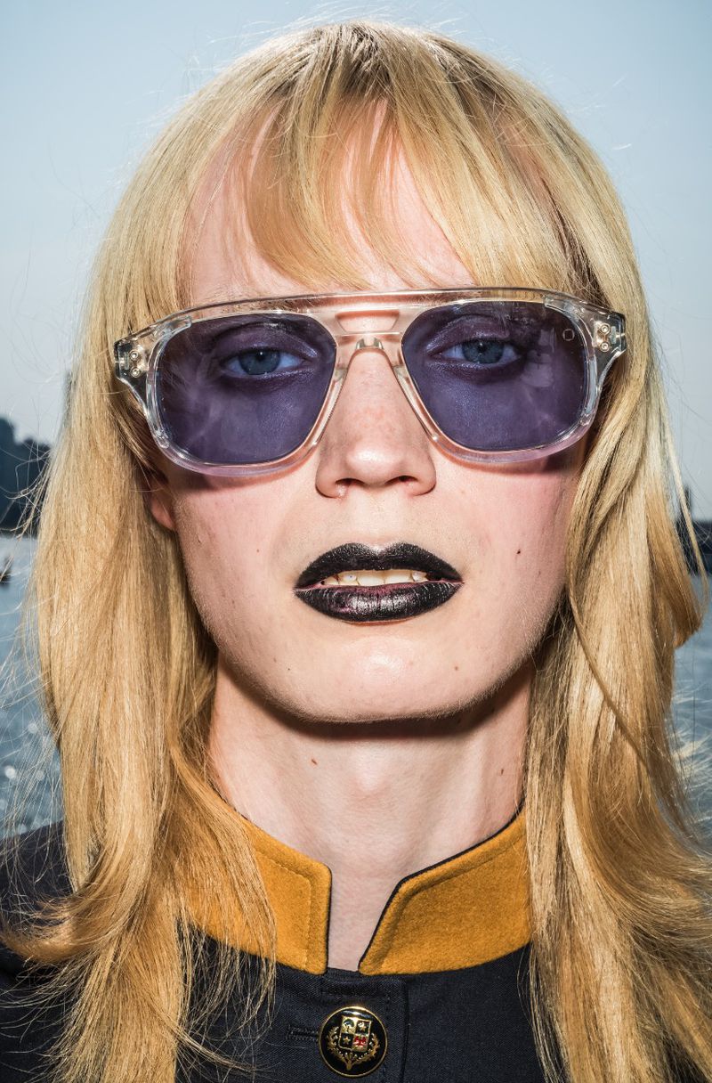 Close-up portrait of a blonde woman with sunglasses and dark lipstick against an outdoor water background with buildings in the distance.
