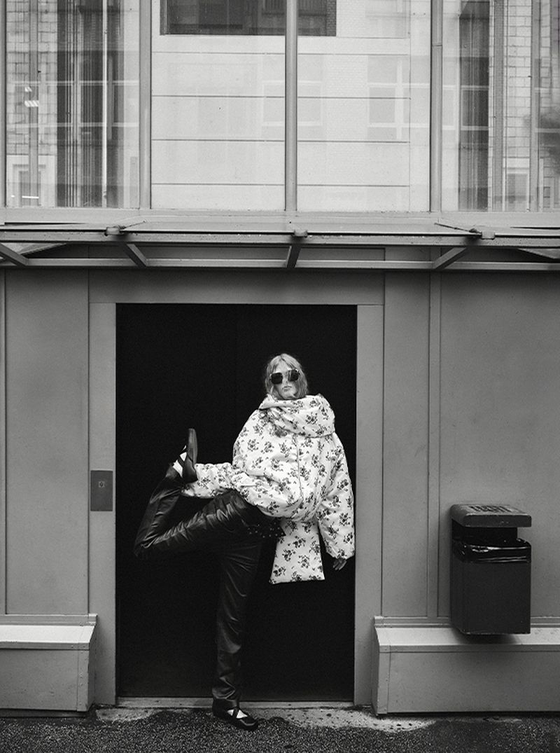 Black and white portrait of a woman wearing a floral jacket and dark sunglasses performing a leg extension in a doorway.