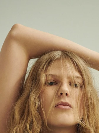 Close-up portrait of a woman with long wavy blonde hair, raising one arm above her head against a plain light background.
