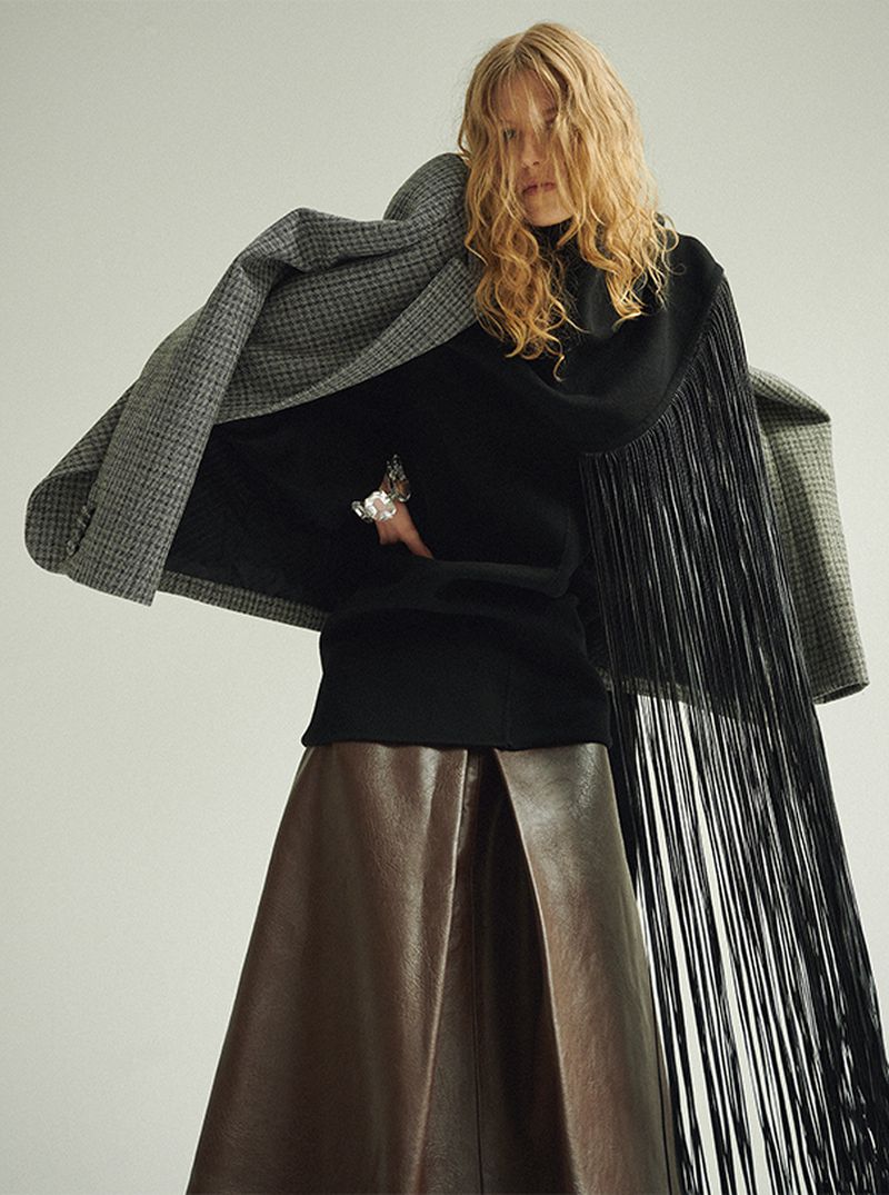 A woman in a black turtleneck sweater, long brown skirt, grey jacket, and black fringe stands against a white background in a studio shot.