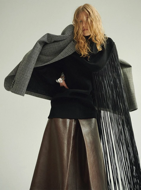 A woman in a black turtleneck sweater, long brown skirt, grey jacket, and black fringe stands against a white background in a studio shot.