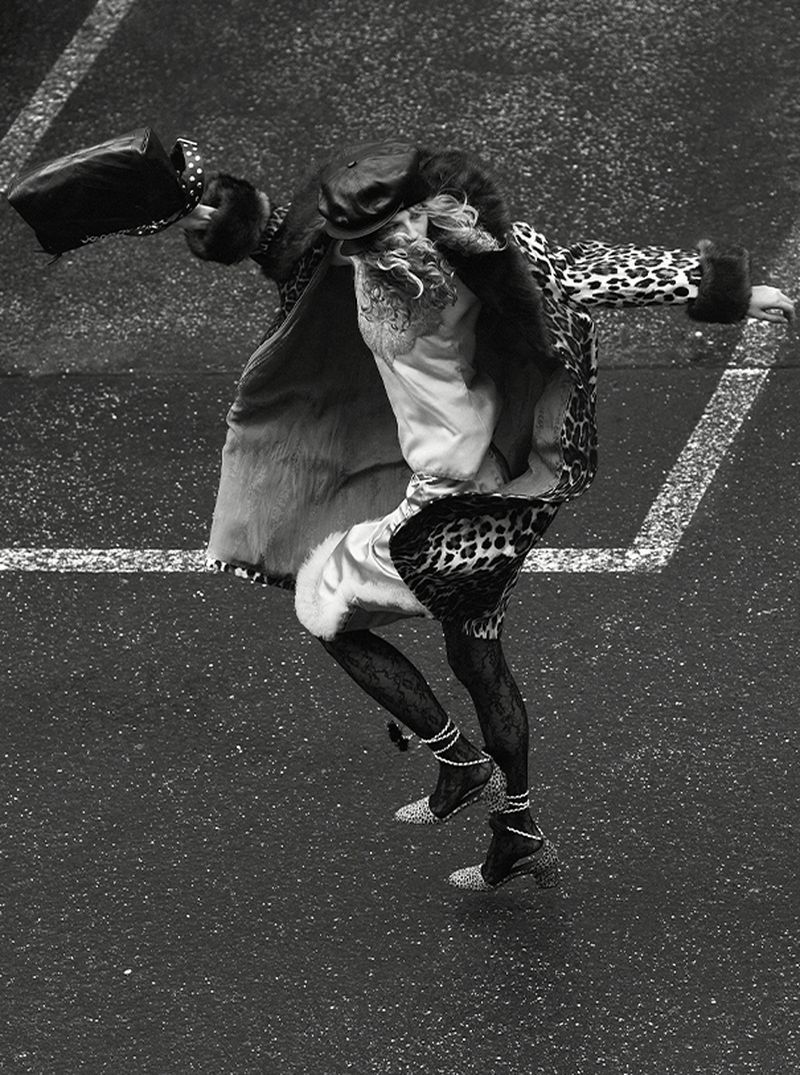 Fashionable individual in an animal print coat, tights, and heels, balancing a bag, captured in a high-angle shot on a paved surface.