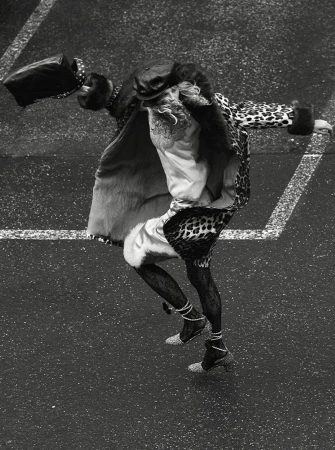 Fashionable individual in an animal print coat, tights, and heels, balancing a bag, captured in a high-angle shot on a paved surface.